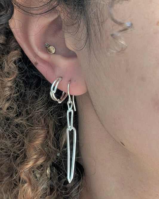 A close-up of a person's ear wearing a pair of silver hoop earrings with long, thin dangling bar elements, alongside other ear piercings.