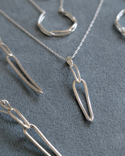 A close-up of a silver necklace with a minimalist, open geometric pendant on a textured blue background, with other matching silver jewelry pieces partially visible around it.