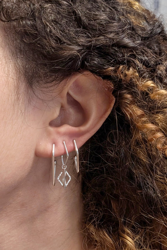 A close-up of a person's ear wearing three silver hoop earrings, including one with a dangling geometric diamond-shaped charm.
