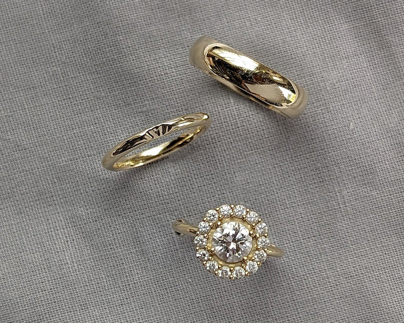 Three yellow gold rings displayed on a textured gray fabric background: a smooth band ring, a twisted band ring, and a halo ring featuring a round clear center stone surrounded by small clear stones.
