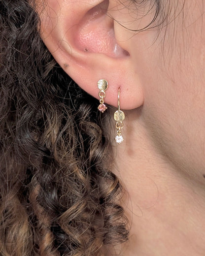 A close-up of a person's ear wearing a delicate gold dangle earring with a small pink gemstone and a clear faceted drop.