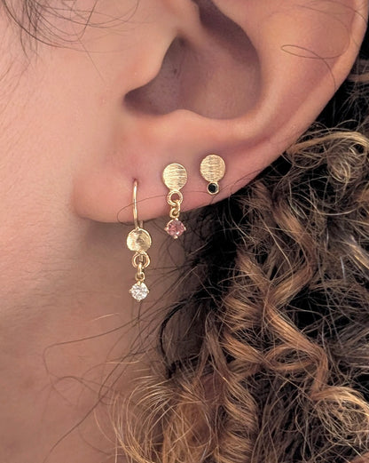 Close-up of a person's ear wearing gold hoop and stud earrings, including textured oval studs, a small pink gemstone drop, and a dangling clear crystal charm.
