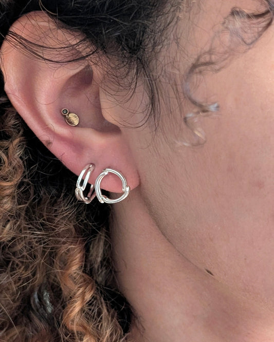 Close-up of a person's ear wearing three hoop earrings: a small gold stud in the cartilage, a silver huggie hoop, and a larger silver hoop with a twisted front detail.
