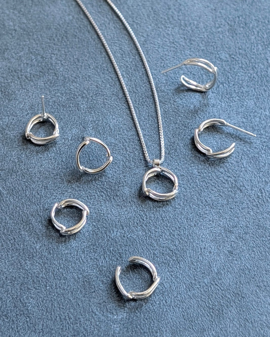A jewelry set featuring silver ring spacer charm, ear cuff, pendant necklace on box chain, ring studs and small circle hoops on arranged on a textured blue-gray fabric background.