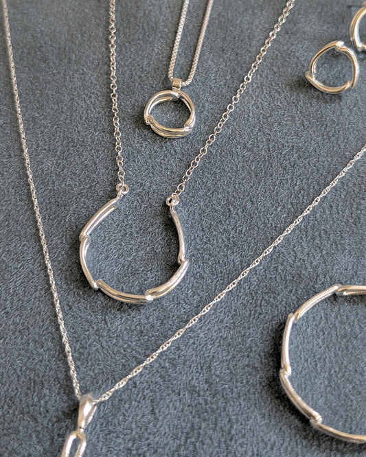 A set of silver jewelry pieces, including a necklace with a segmented circular pendant, a matching hoop earring, and a ring, displayed on a textured blue gray background.
