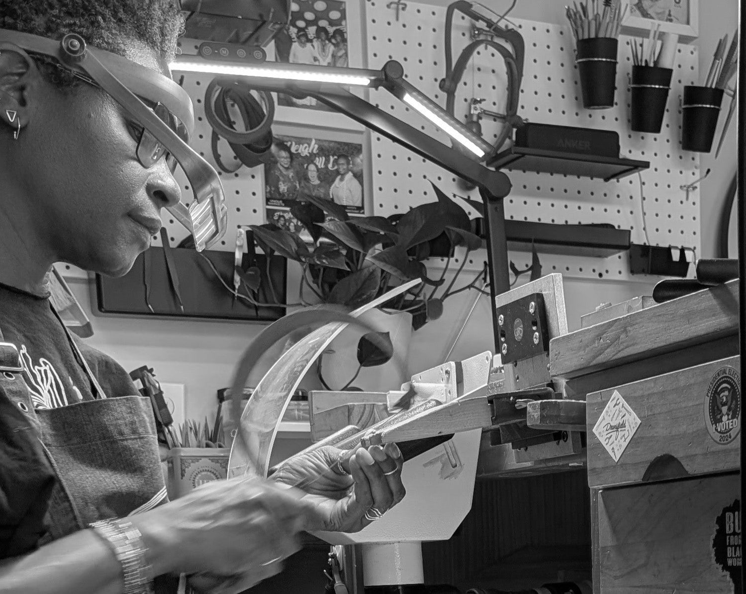 A black and white photograph of a craftsman wearing protective glasses and an apron, using a woodworking machine in a workshop with a pegboard background.