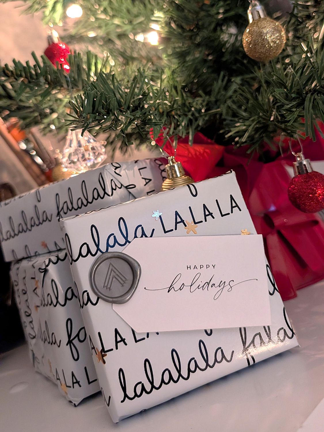 Gift wrapped in 'lalala' paper with a 'Happy Holidays' card, Christmas tree in the background.