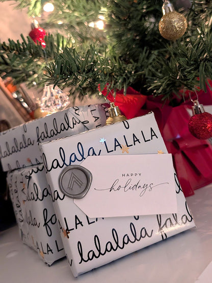 Gift wrapped in 'lalala' paper with a 'Happy Holidays' card, Christmas tree in the background.