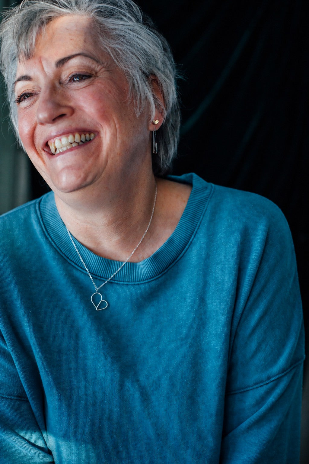 Joyful model wearing heart necklace and earrings wearing a blue shirt.