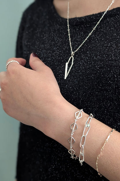 A close-up of a person wearing multiple silver-toned jewelry pieces, including a long necklace with a geometric pendant, a ring, and several bracelets on the wrist, against a black sparkly top and a pale blue background.