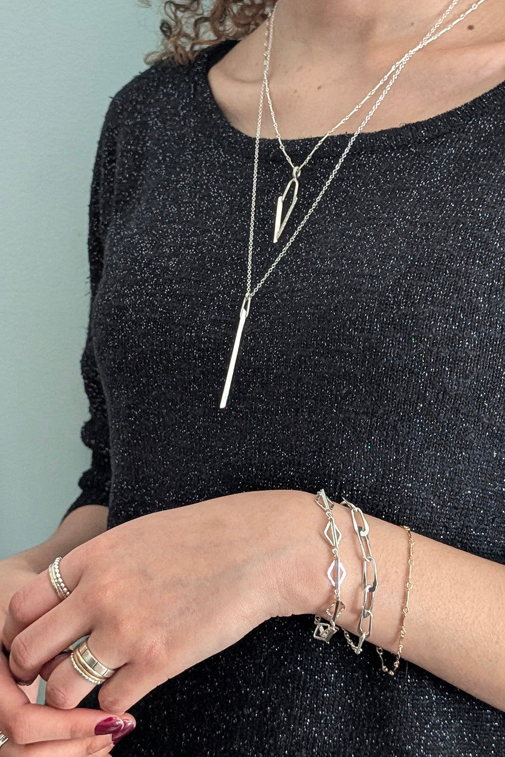 A close-up of a person wearing layered silver necklaces, bracelets, and rings, with a long vertical bar pendant necklace and geometric chain pieces. The person is wearing a sparkly black top against a pale blue-gray background.