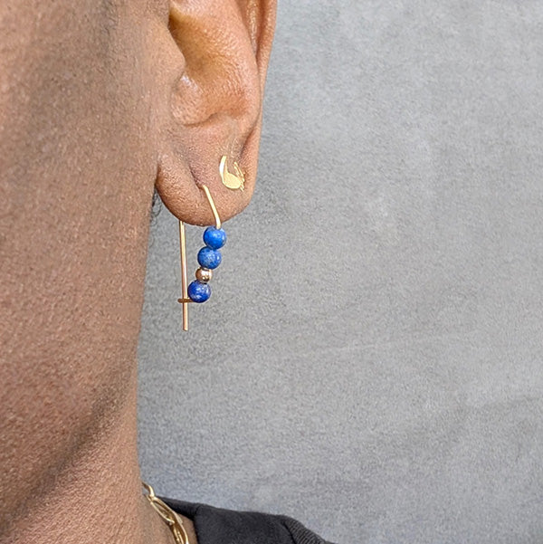 Close-up of an ear wearing a gold earring with blue beads against a gray background