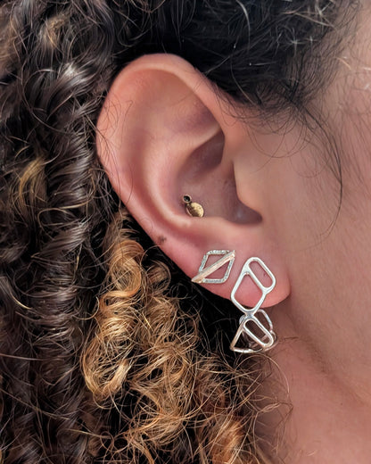 A close-up of a person's ear showcasing a pair of silver geometric hoop earrings on the lobe, with a small gold stud on the helix.