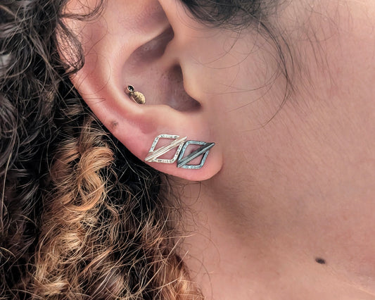 A close-up of a person's ear wearing a pair of geometric silver stud earrings with a textured, hammered finish. The person has light skin and curly brown hair.
