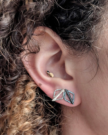 A close-up of a person's ear showcasing a silver geometric stud earring with diamond-shaped facets, alongside other ear piercings.