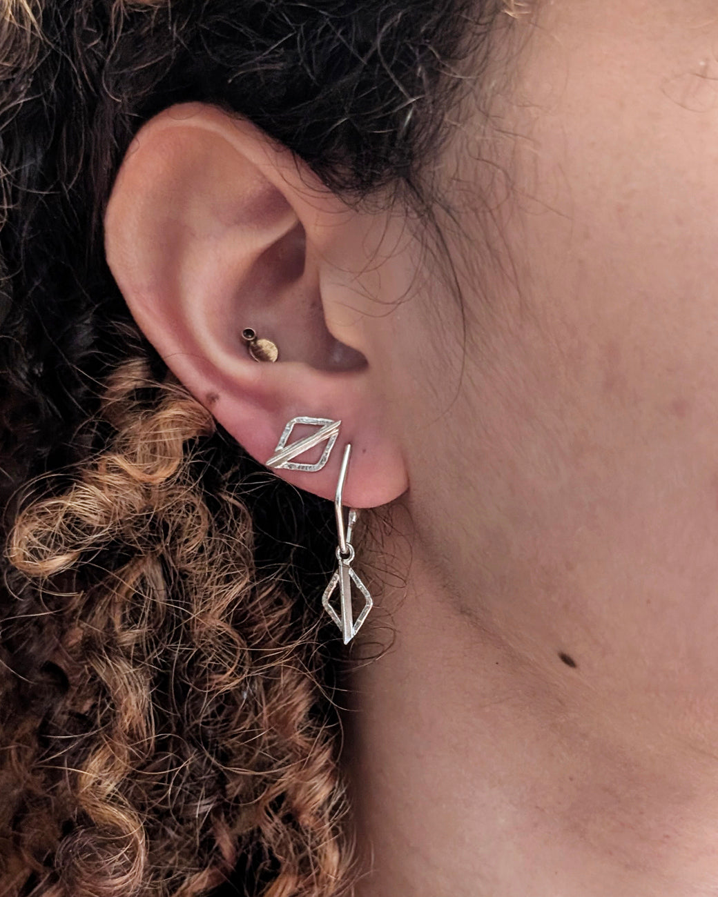A close-up of a person's ear showcasing a silver geometric dangle earring with a small diamond-shaped drop and a matching geometric stud, alongside other ear piercings.