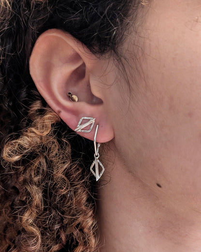 A close-up of a person's ear showcasing a silver geometric dangle earring with a small diamond-shaped drop and a matching geometric stud, alongside other ear piercings.
